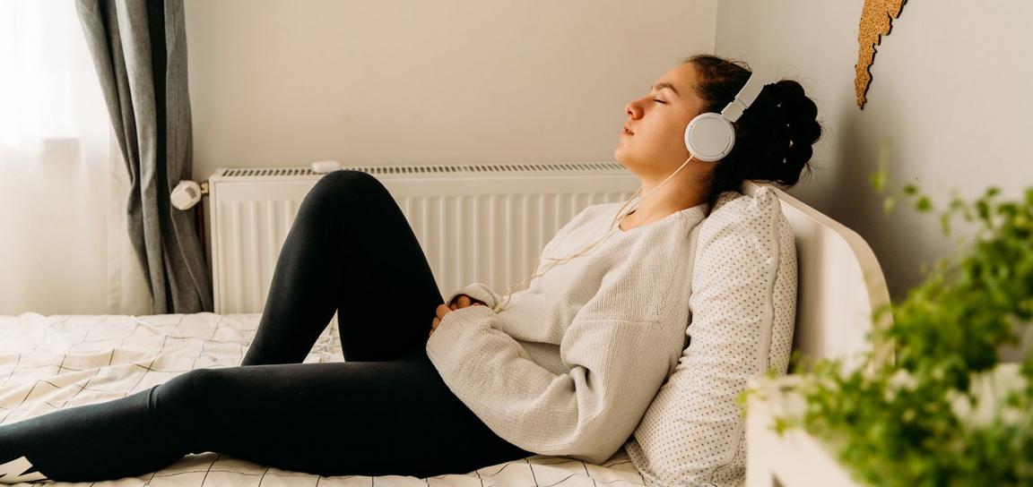 A person with white headphones wearing a white jumper and black leggings sat up on a bed