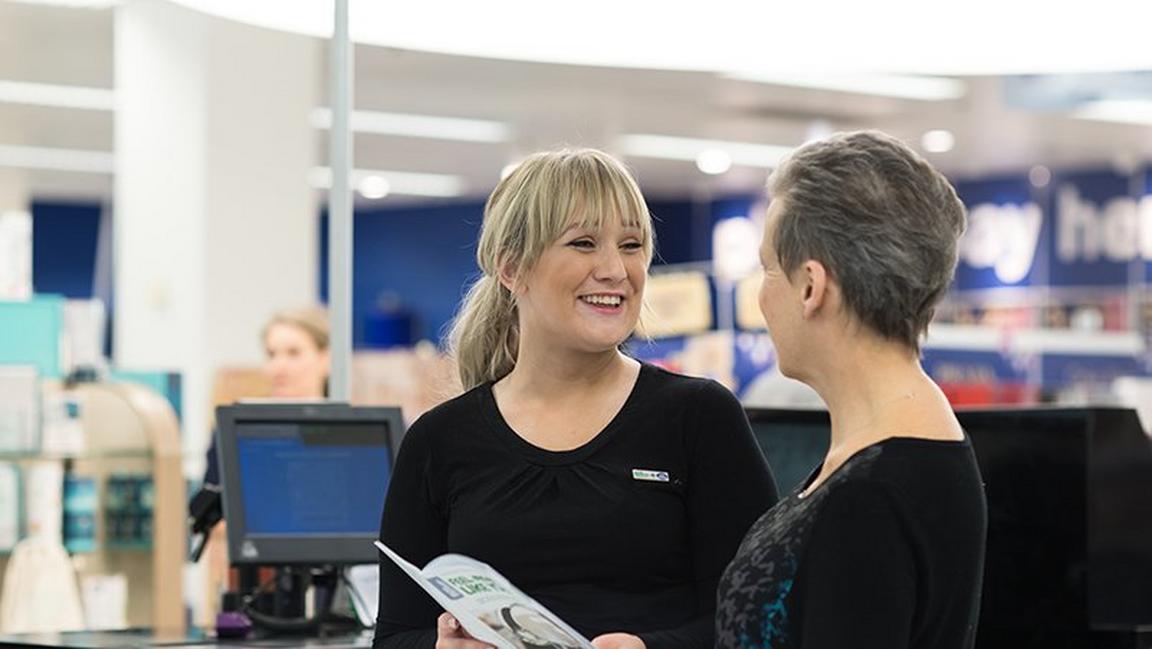 Two women talking and smiling in a retail environment, with one holding a brochure.