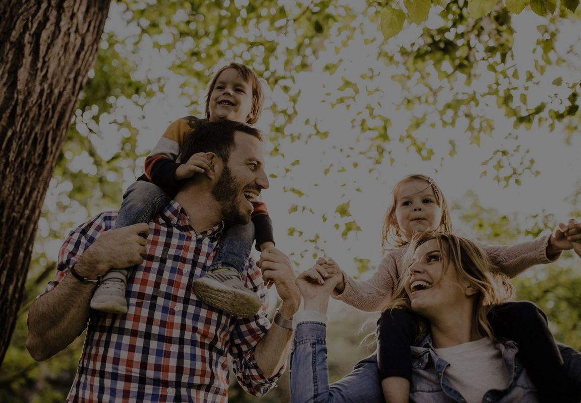 A family of four enjoying a sunny day in a leafy area; two adults carrying children on their shoulders.