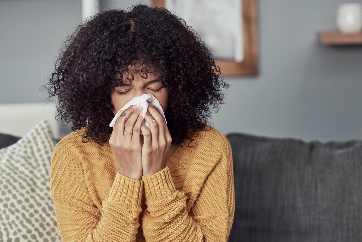 Person with curly hair in a yellow sweater holding a tissue to their nose on a gray sofa.