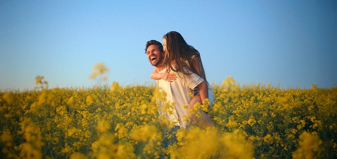 A man giving a piggyback ride to a woman in a field of yellow flowers under a blue sky.