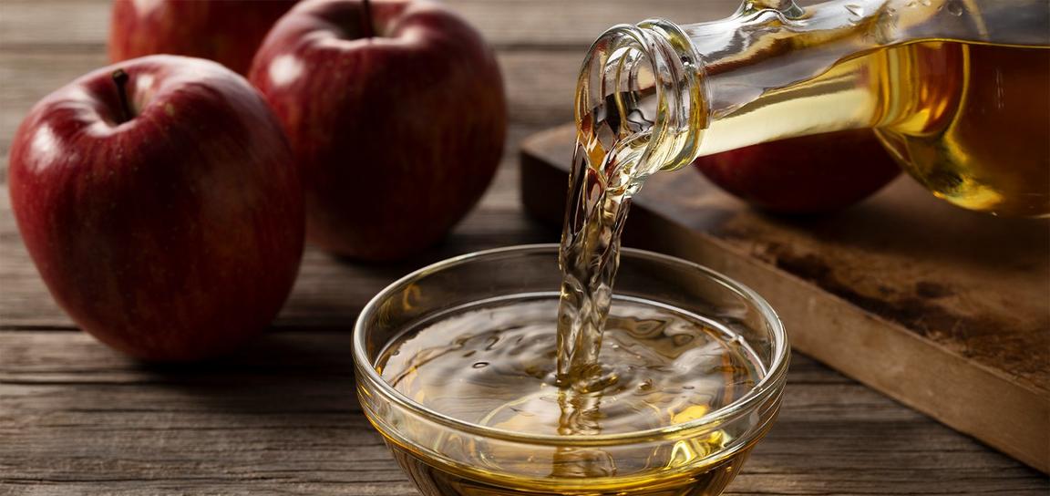 Apple cider being poured into a glass bowl with red apples in the background.