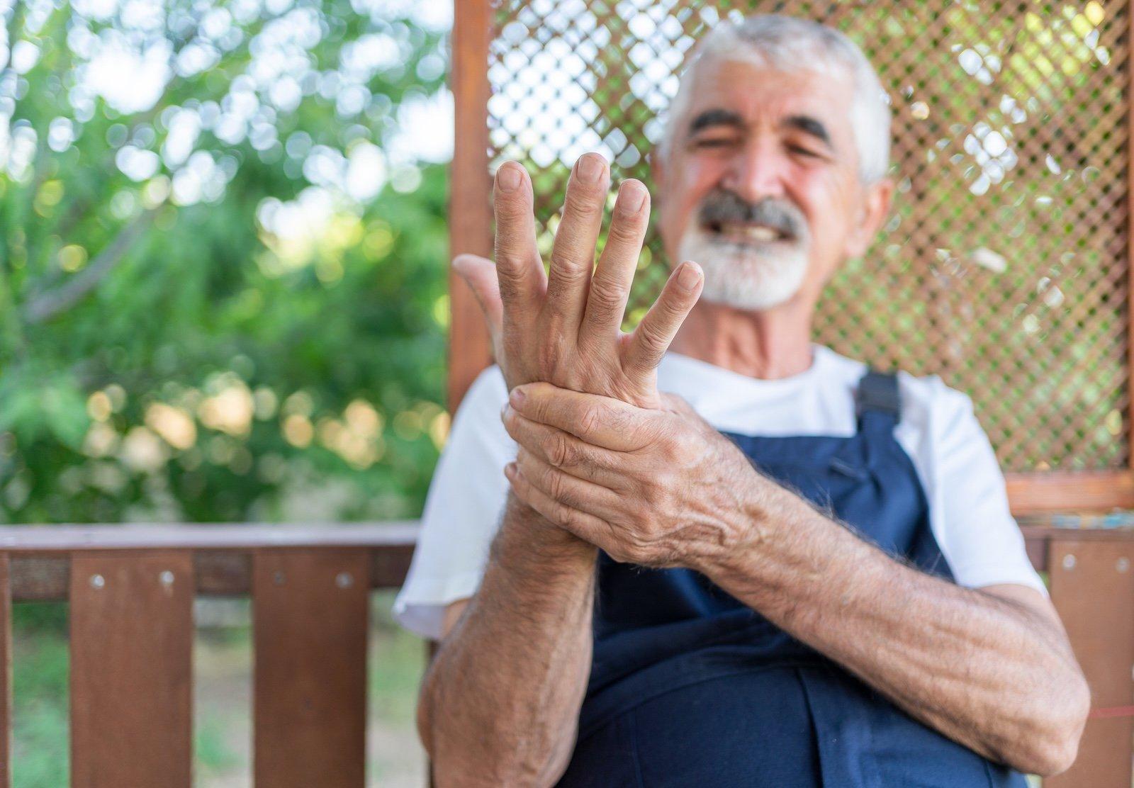Older man holding his hand in pain while sitting outdoors on a wooden bench with greenery in the background