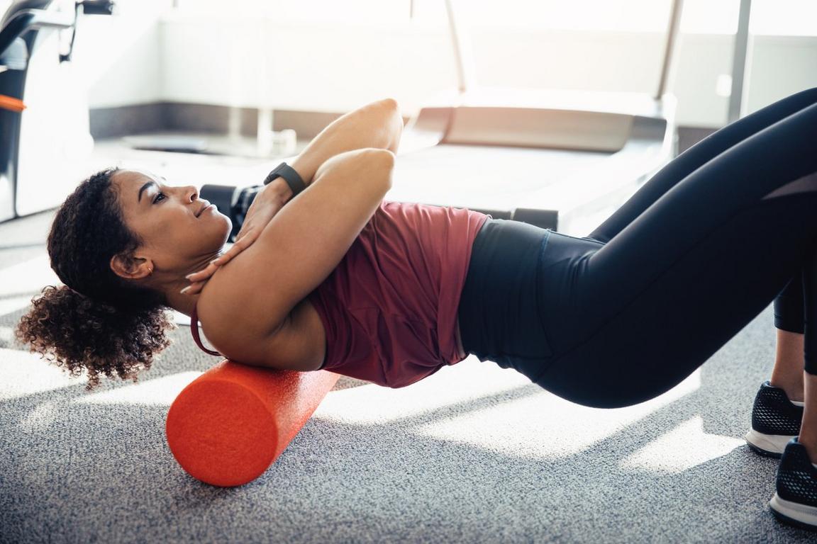 A person using an orange foam roller on a gray carpeted floor.