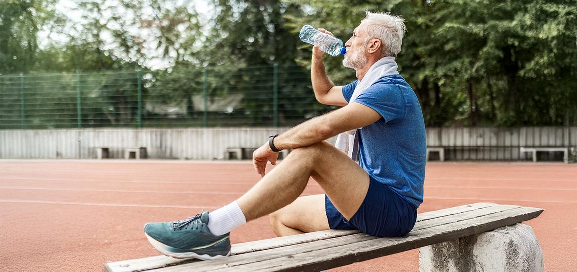 Older man in athletic wear sitting on a bench drinking water beside a running track.