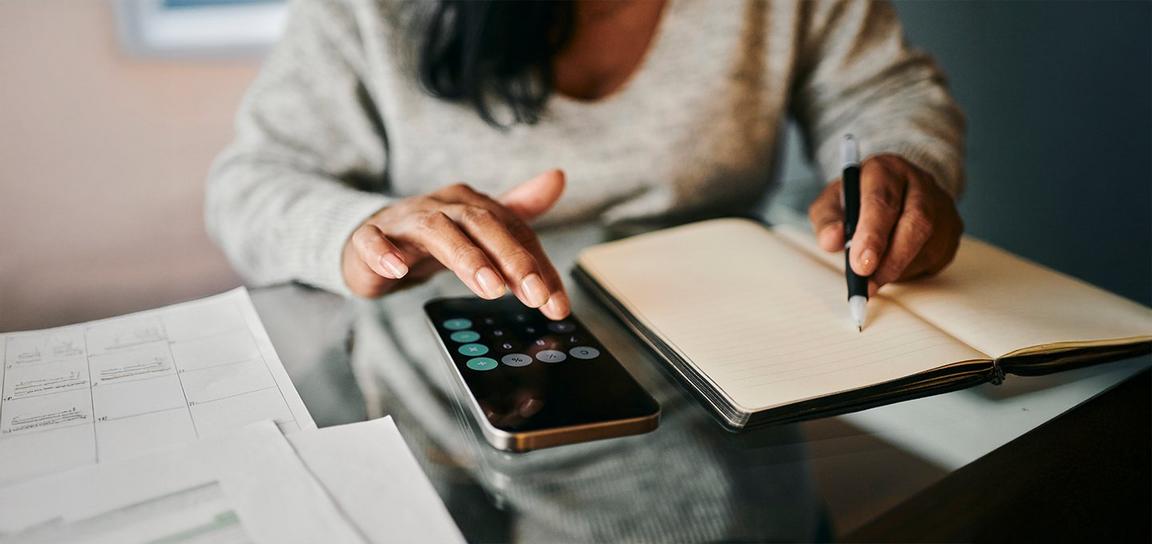 Person using a smartphone calculator and writing in a notebook at a glass table.