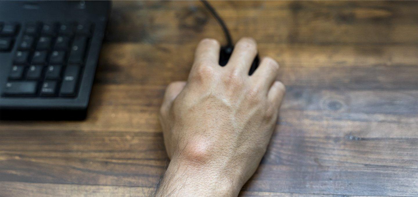 A hand using a black computer mouse on a wooden desk, with part of a keyboard visible.