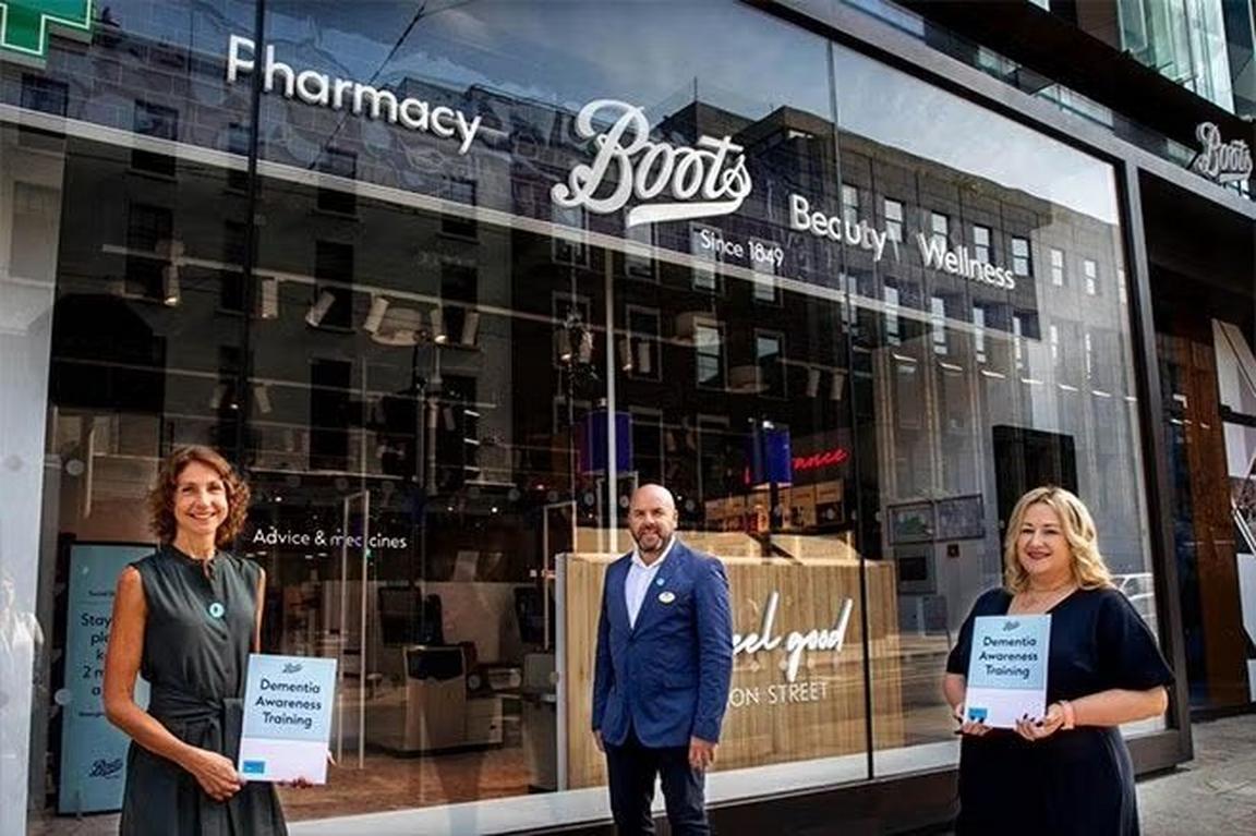 Three people stand outside a Boots pharmacy, holding certificates for Dementia Awareness Training.