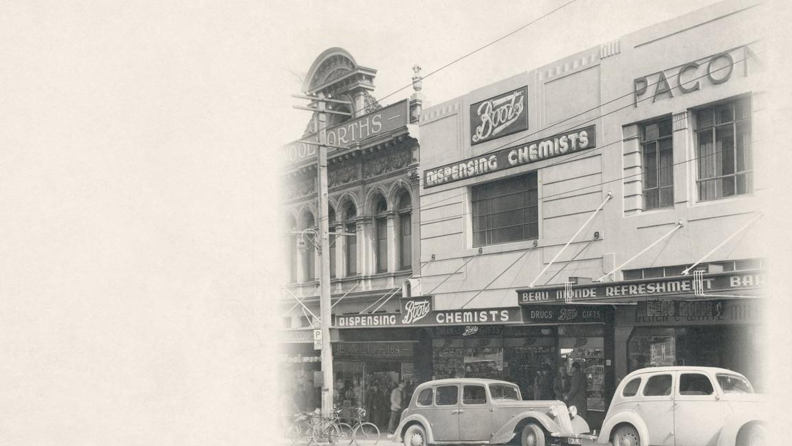 Historic street scene with vintage cars and buildings labeled "Woolworths" and "Boots Dispensing Chemists.