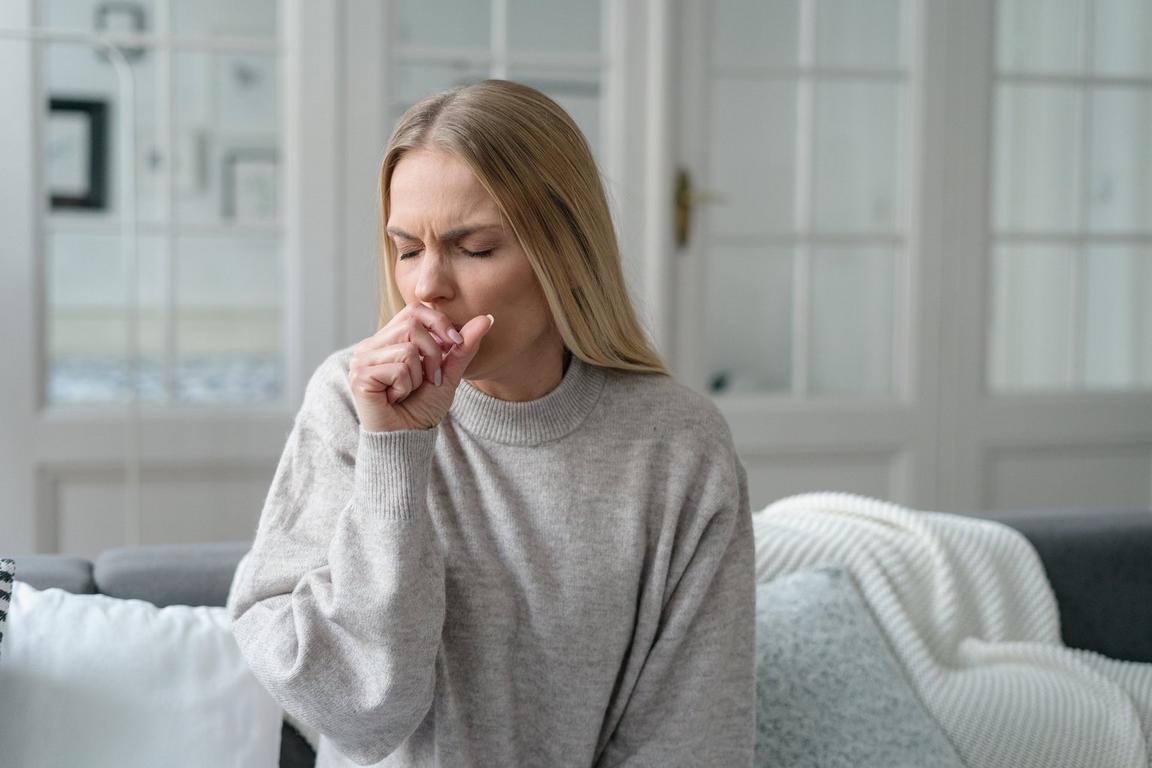 Person in a gray sweater sitting on a sofa, covering their mouth as they cough.