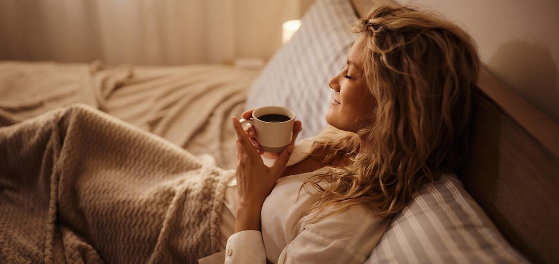 A woman holding a mug while relaxing in bed with a beige blanket and striped pillows.