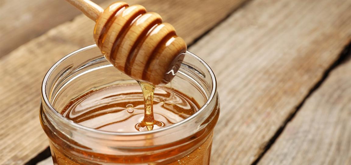 A honey dipper with honey above a jar filled with honey on a wooden surface.