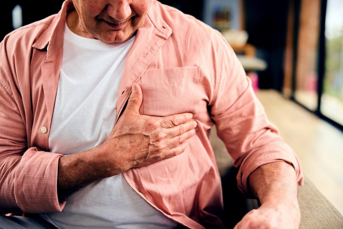 Man in a salmon shirt holding his chest while seated indoors.