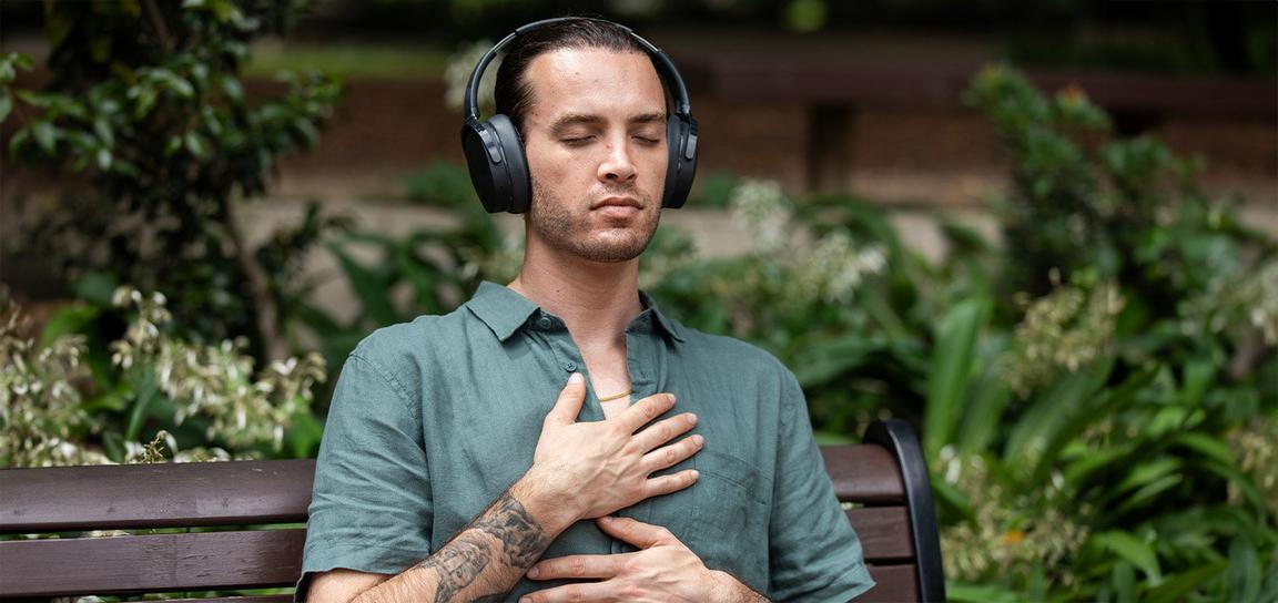 A person with headphones sitting on a bench, eyes closed, hand on chest, in a garden.