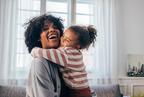 A woman laughing as she is hugged by a smiling young girl indoors, with a window and curtains in the background