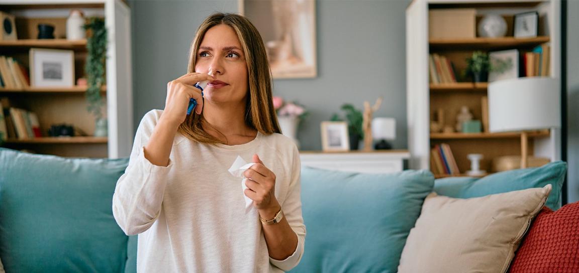 Woman holding nasal spray and tissue on a couch in a living room.