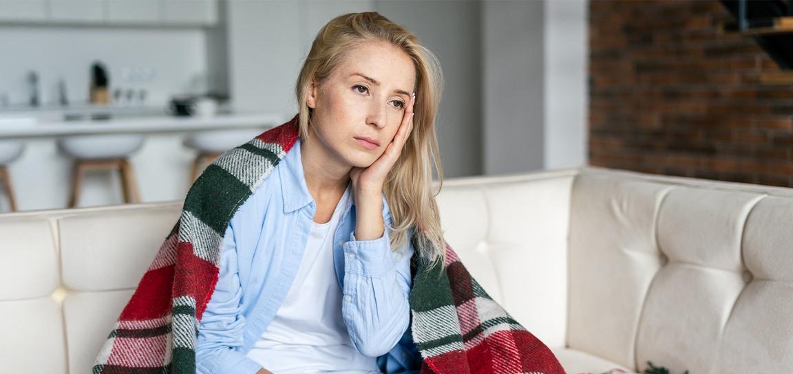 A person sitting on a sofa with a plaid blanket over their shoulders, looking contemplative.