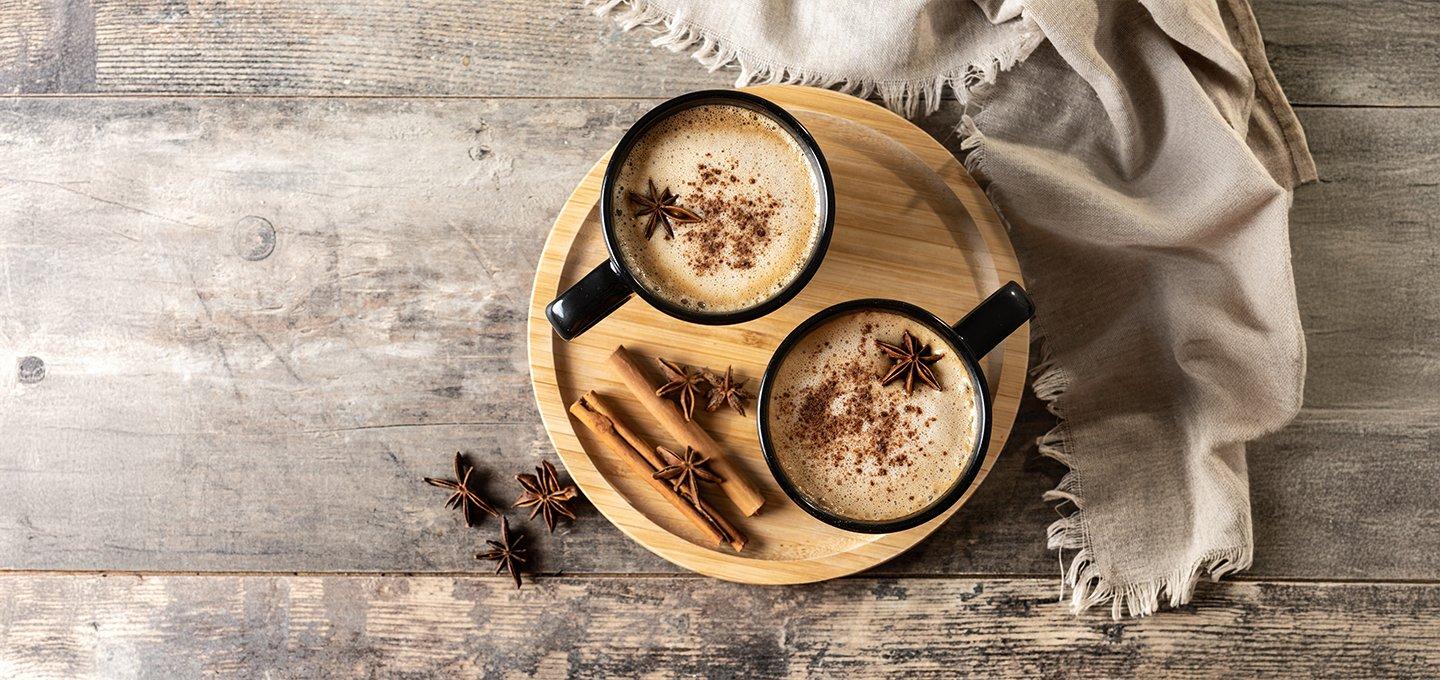 Two mugs of frothy coffee on a wooden tray with cinnamon and star anise.