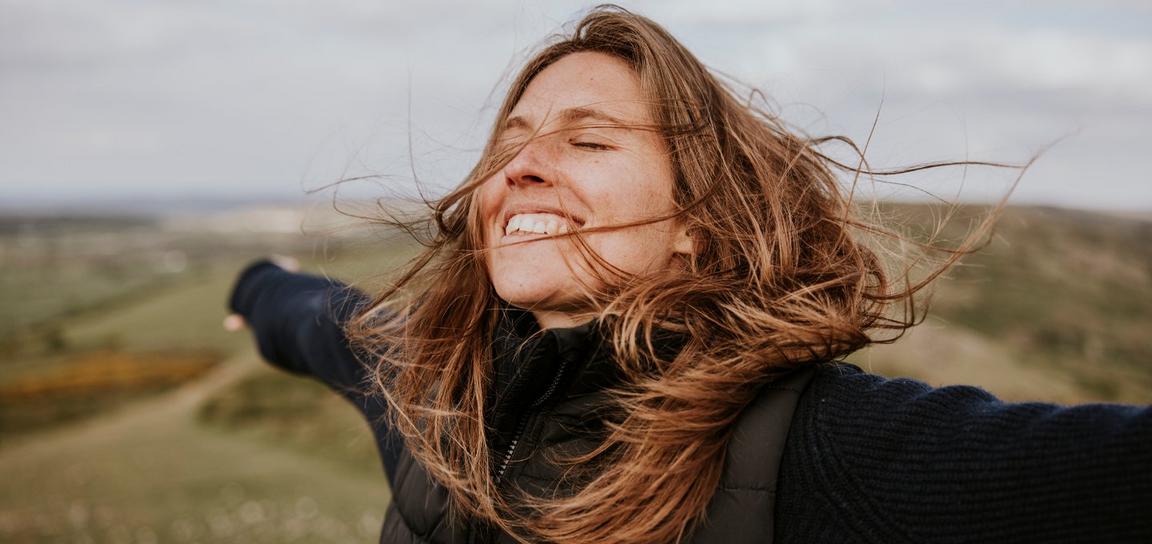 A woman with brown hair and a smile stands outdoors with her arms spread wide, set against green rolling hills and a cloudy sky.