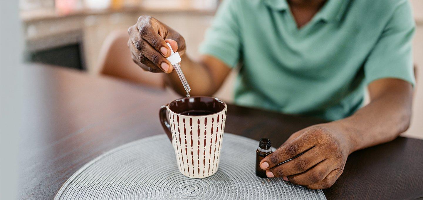 Person adding drops of liquid from a dropper bottle into a mug on a table.