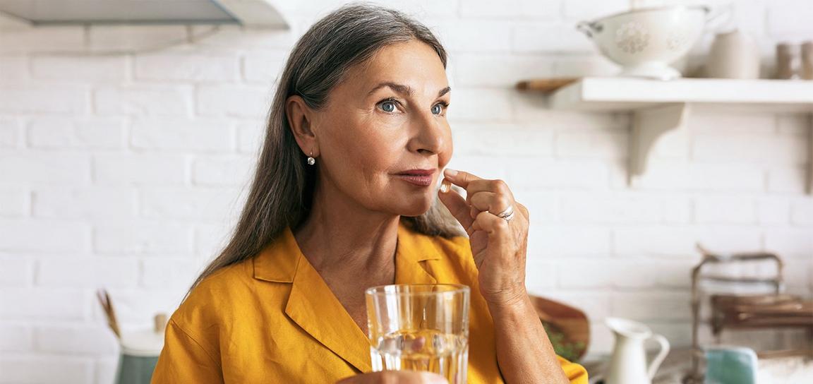 Woman holding a pill and a glass of water in a kitchen setting.
