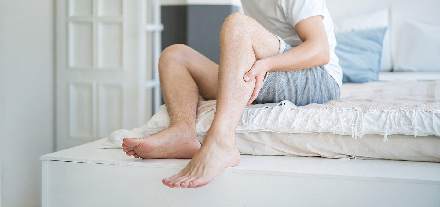 Person sitting on the edge of a bed, holding their lower leg in a bright bedroom.