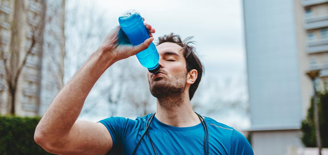 Person drinking from a blue water bottle outdoors with blurred buildings in the background.