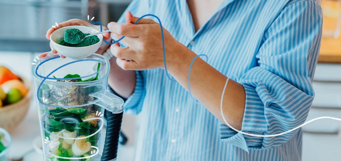A person in a striped shirt adds green powder to a blender with spinach and seeds.