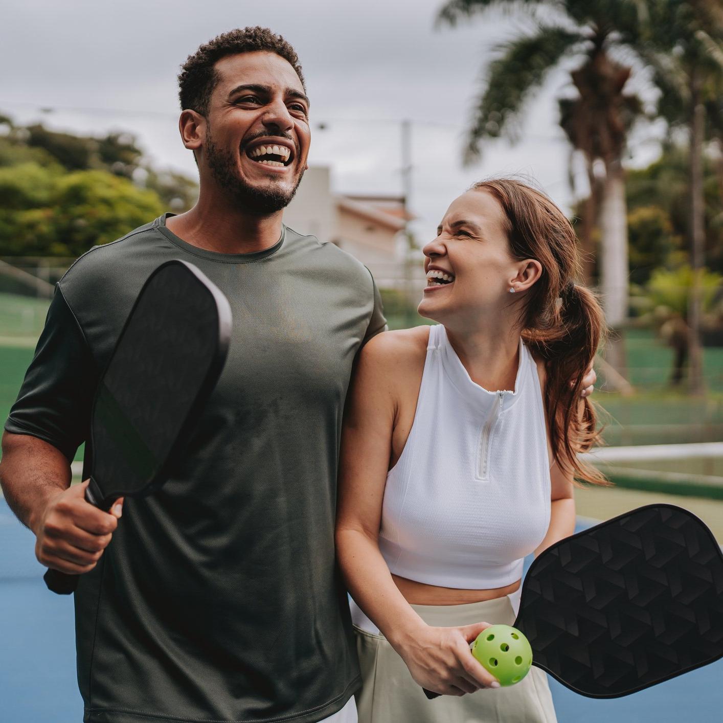 A man and woman smiling on a sports court, holding paddles and a pickleball.