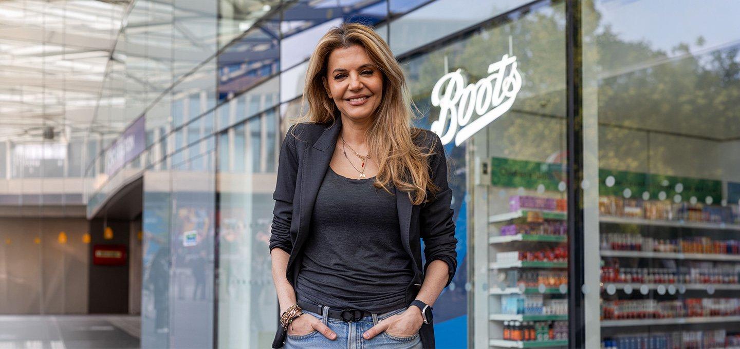 Person standing outside a Boots store, wearing a dark blazer, dark top, and jeans, with hands in pockets.