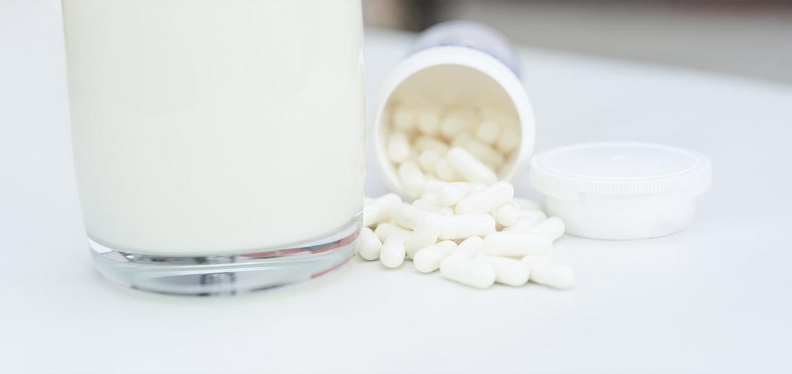A glass of white liquid next to a tipped-over container with white capsules spilling out.