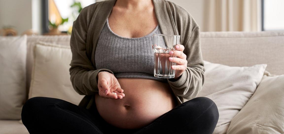Pregnant person holding a pill and a glass of water while sitting on a sofa.