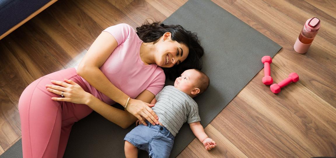 A woman lying on a yoga mat smiling at a baby beside her, with dumbbells and a water bottle nearby.
