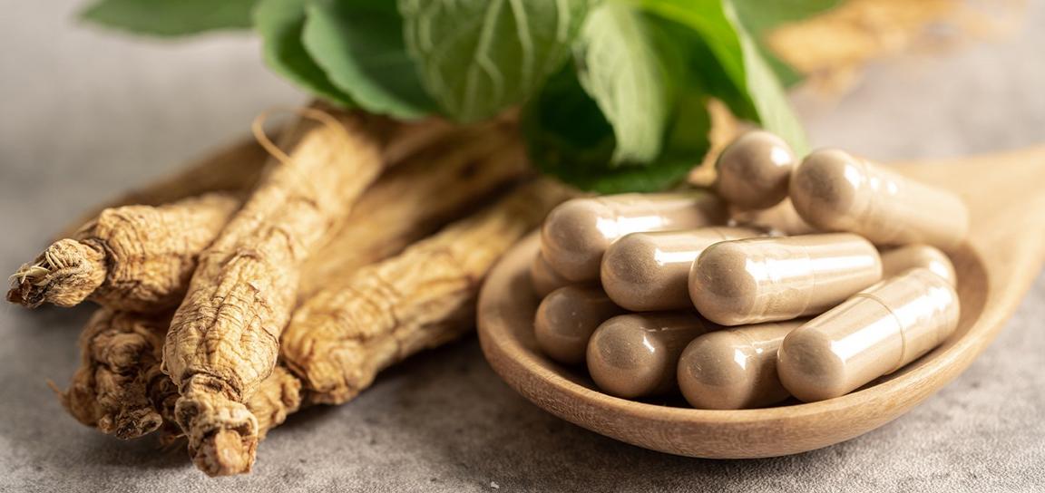 Wooden spoon with beige capsules, dried ginseng roots, and green leaves.