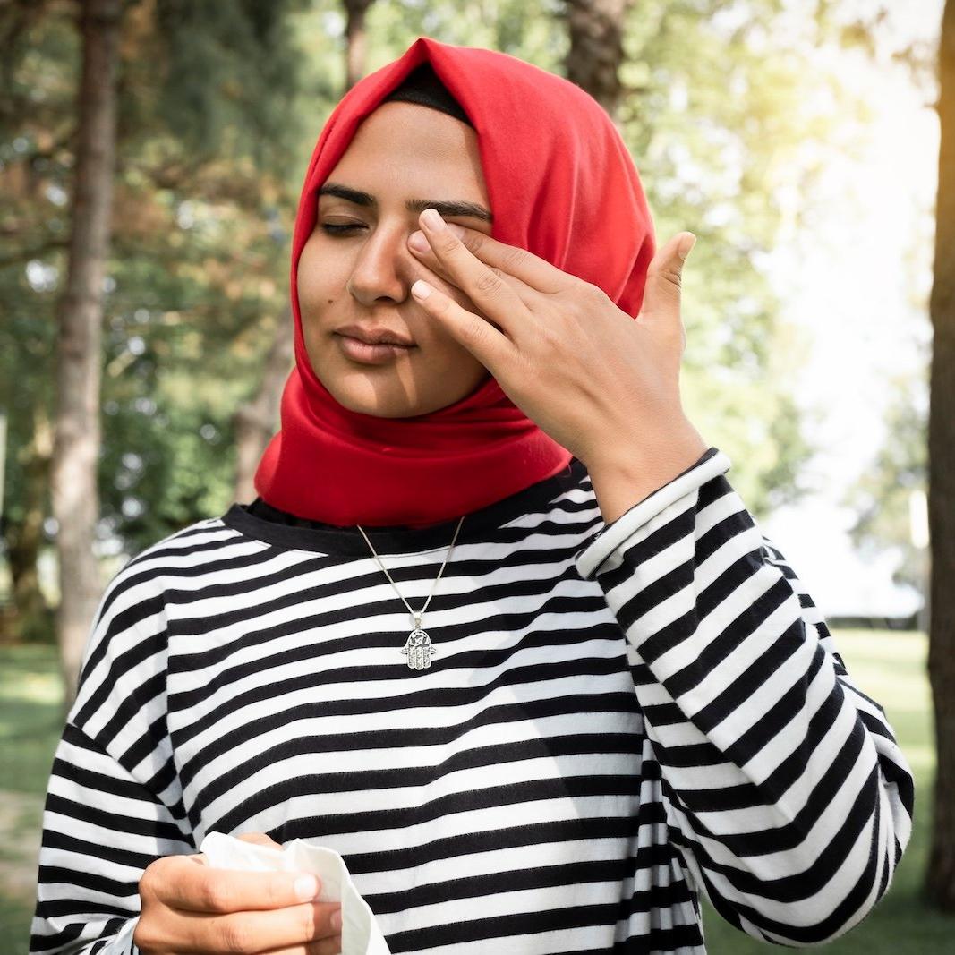 A woman rubbing her eye outside, holding a tissue in the other hand