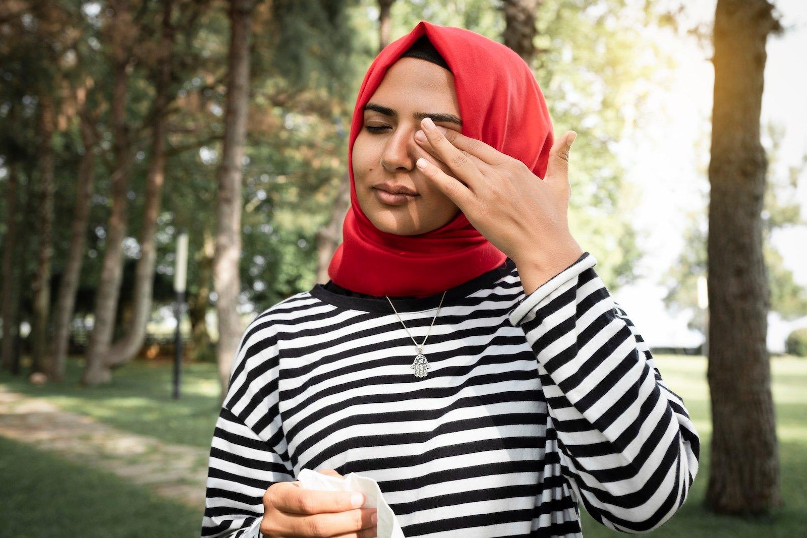 A person rubbing their eye in a forest holding a tissue