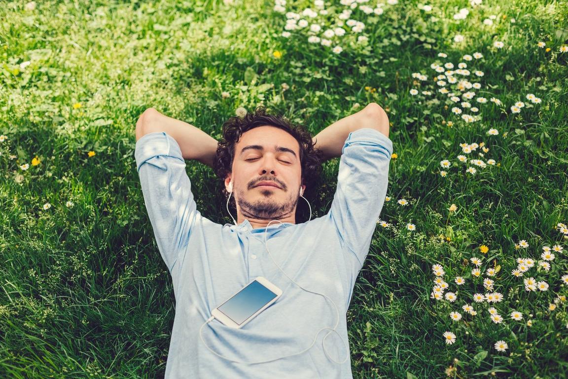 Man relaxing on a green lawn with earphones in, eyes closed, and a smartphone on his chest