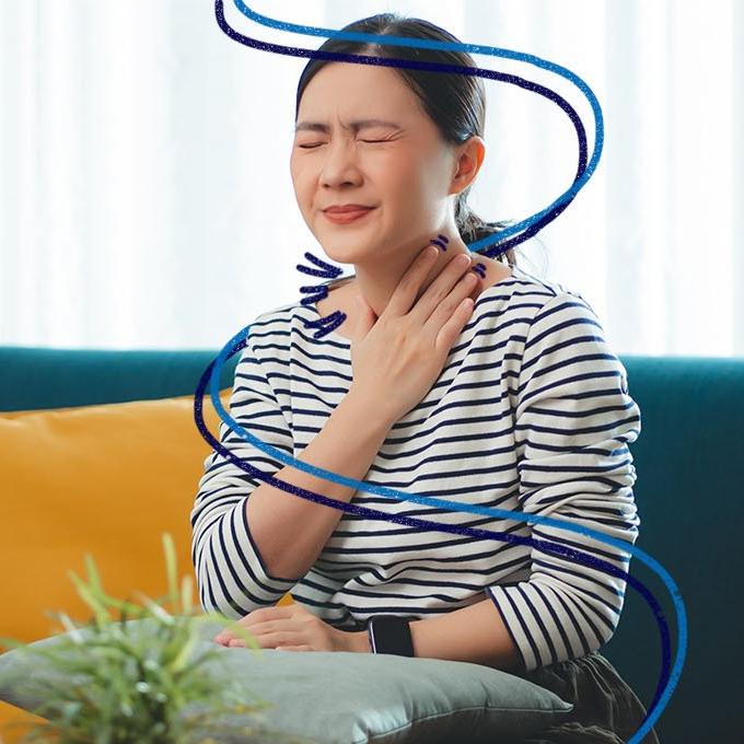 A person sat on a sofa with their eyes closed, holding their throat in pain