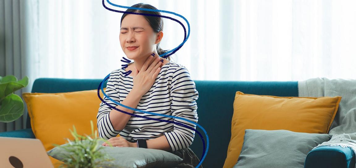 A person sat on a sofa with their eyes closed, holding their throat in pain