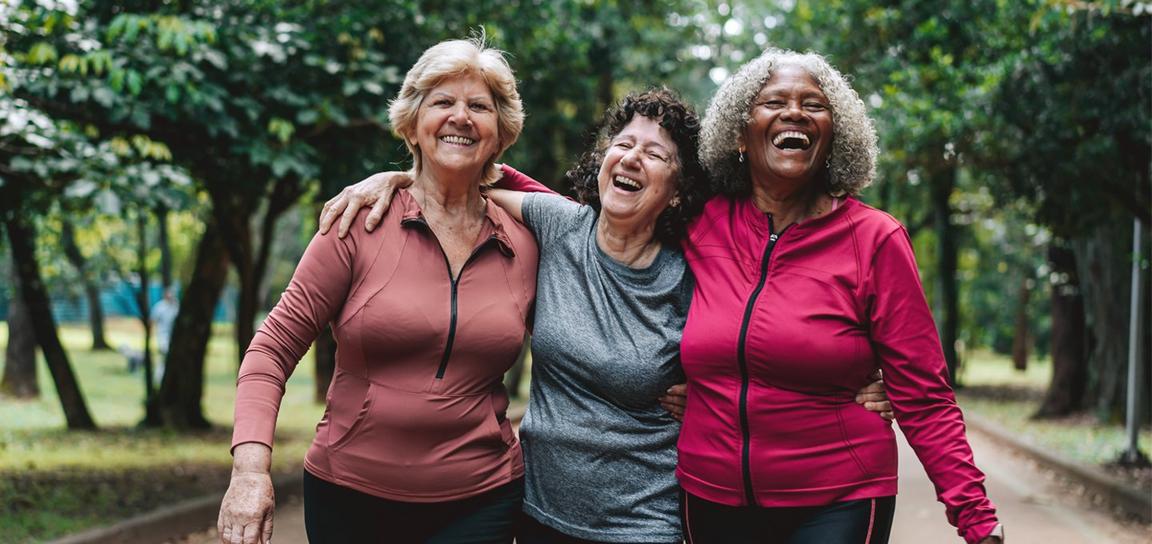 Three women in athletic wear smiling and walking together on a tree-lined pathway, showing joy and friendship.