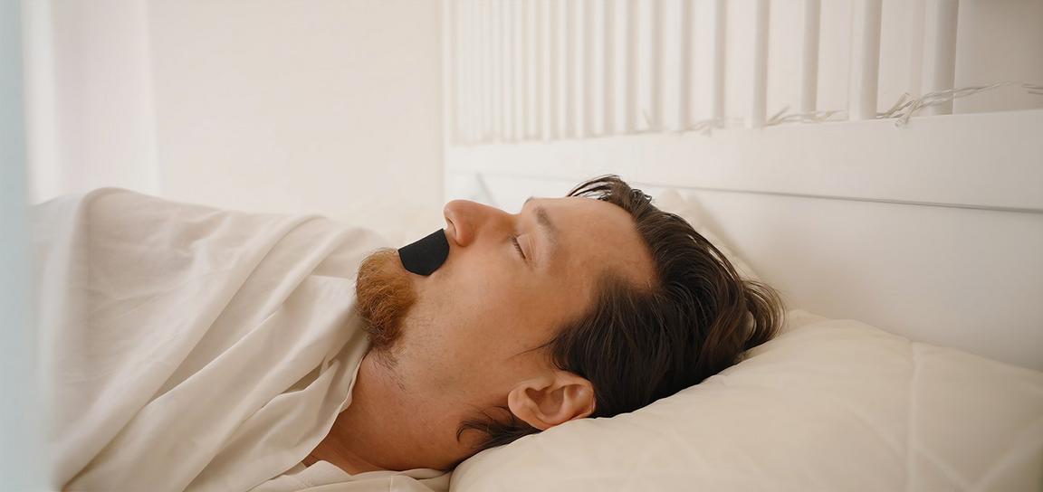 A man with a black tape over his mouth sleeping in a white bed.