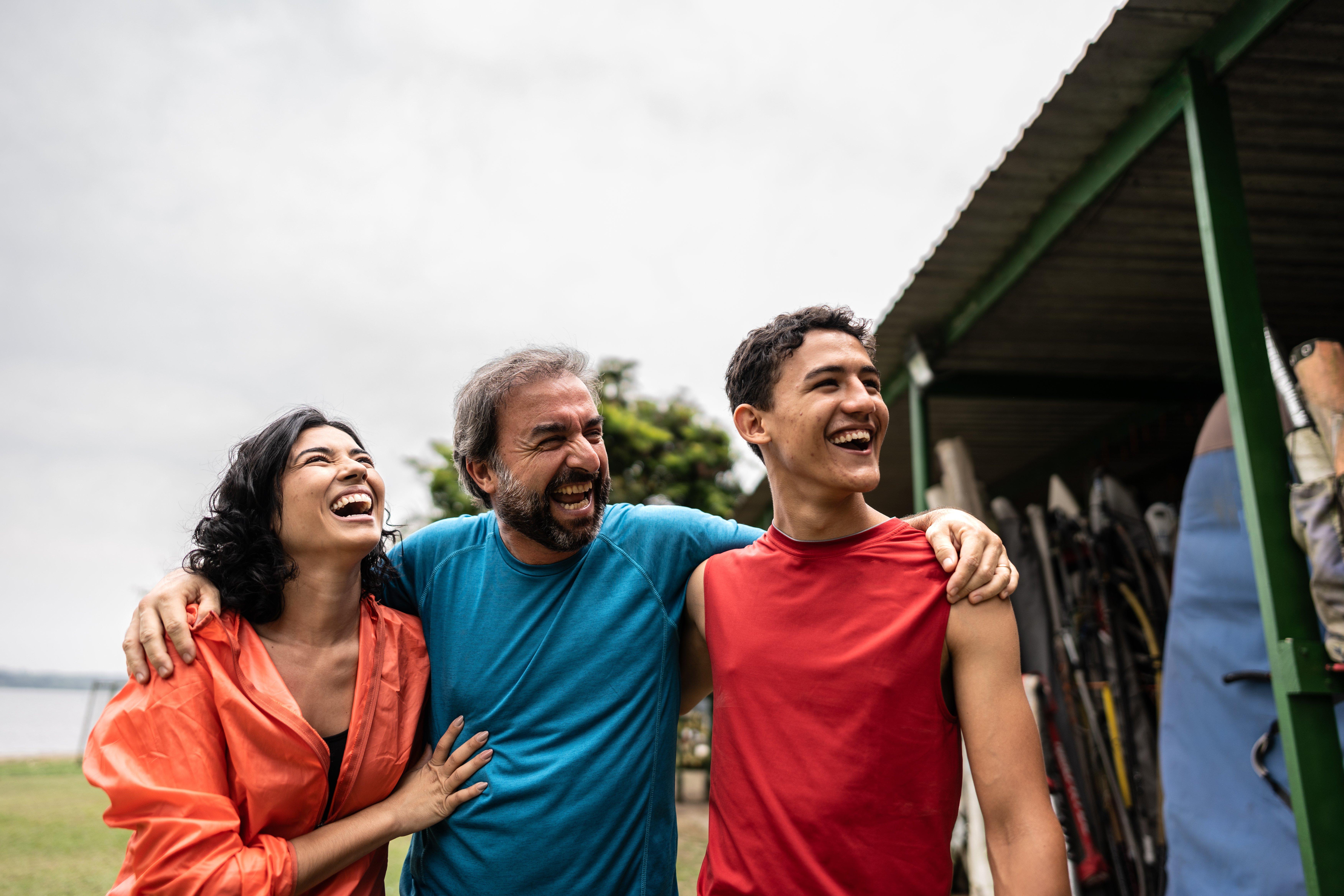 Three people laughing with arms around each other outdoors near a storage area.