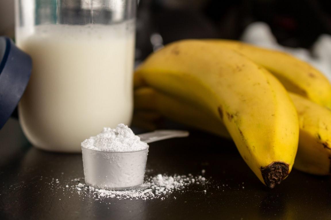 A scoop of white powder on a dark surface, with bananas and a container of liquid in the background.