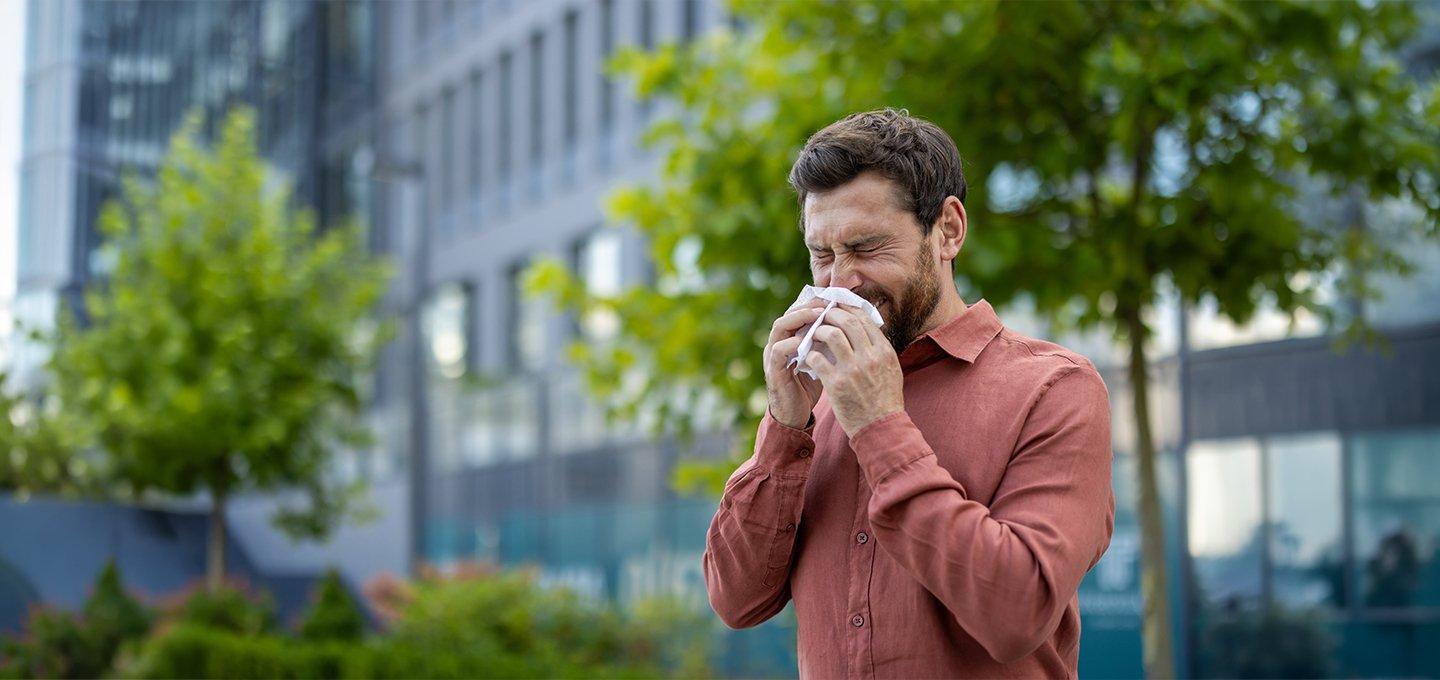 A man in a brown shirt sneezes into a tissue while standing in an urban setting with trees and modern buildings in the background.