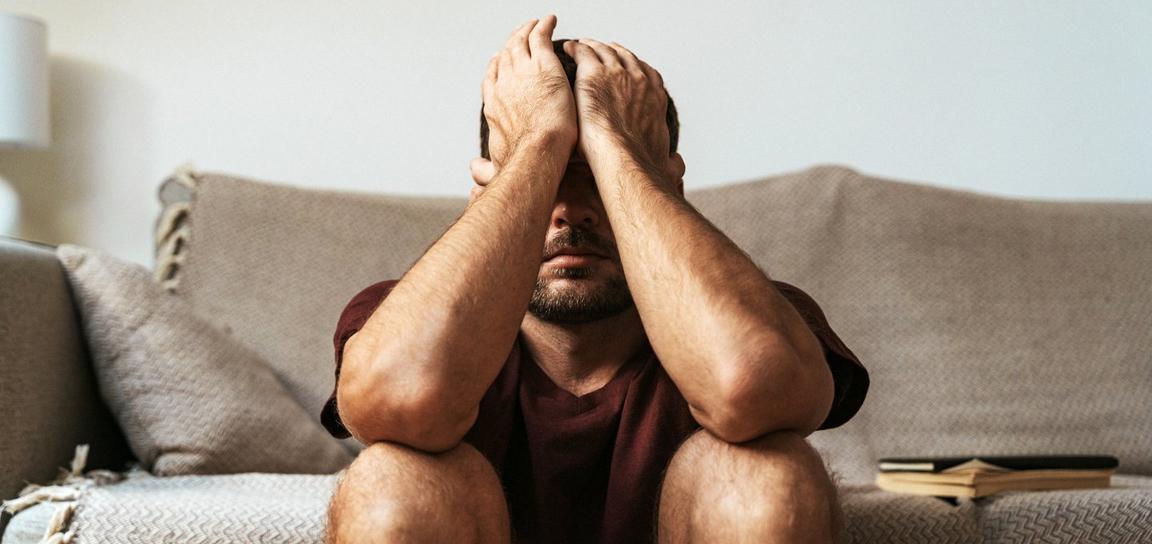 Somebody sat on the floor, against a sofa with their hands over their face