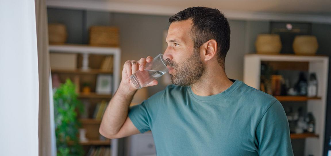 A man in a teal t-shirt drinks water from a glass indoors.