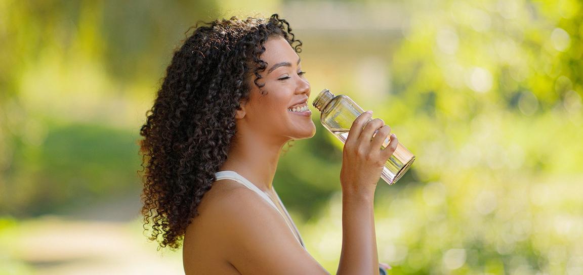 A person smiling while holding a clear water bottle in a green, outdoor setting.