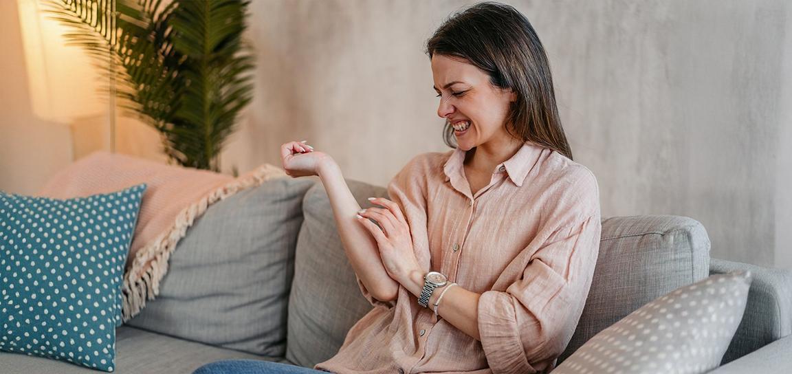 Woman smiling and looking at her arm while seated on a gray sofa with decorative pillows, a plant, and a floor lamp in the background.