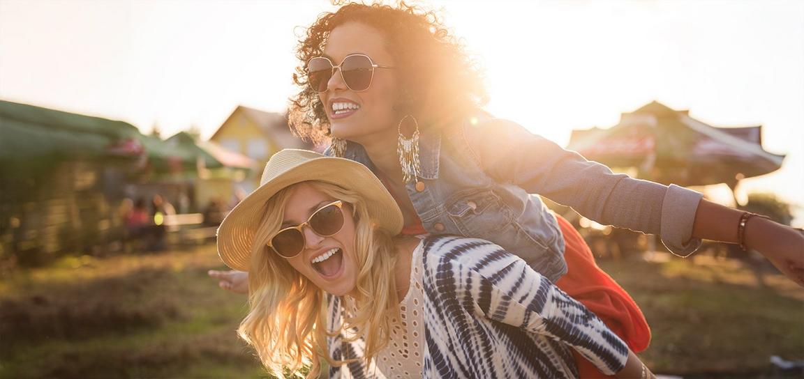 Two women having fun, with one giving the other a piggyback ride in a sunny outdoor setting.