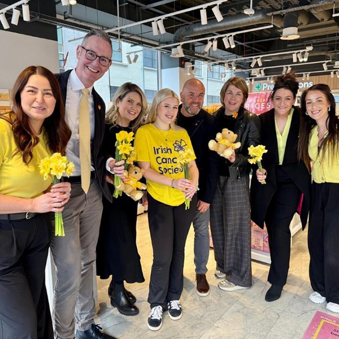 A group of eight people holding yellow daffodils and teddy bears, with some wearing yellow clothing.